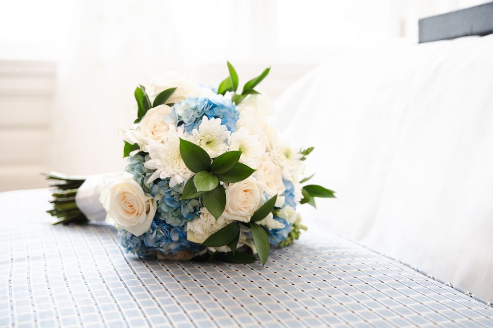 A bridal bouquet of white roses and blue hydrangeas with green leaves resting on a white quilted surface.