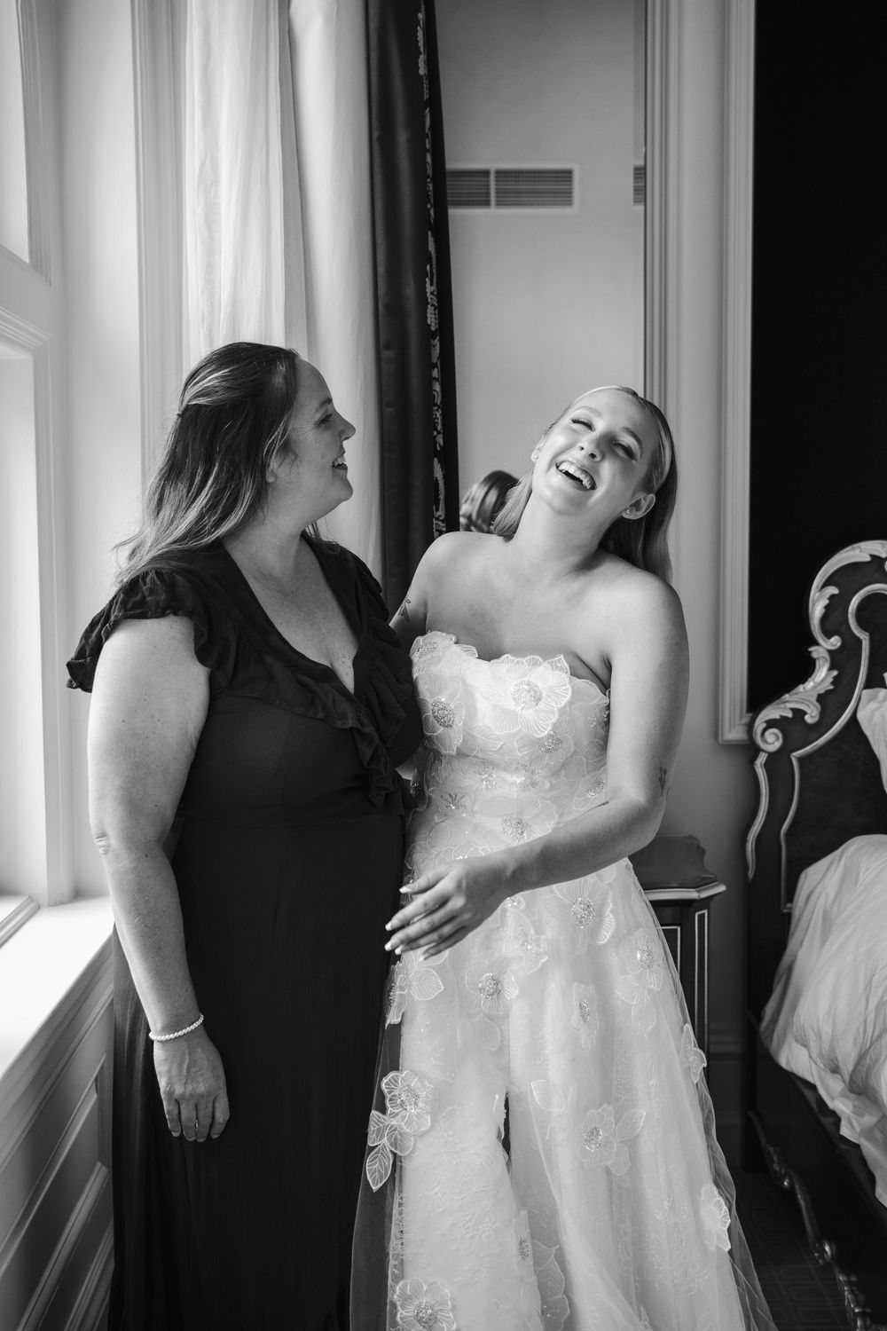 Black and white photo of a laughing bride in a strapless gown with a woman in a dark dress during wedding preparations.