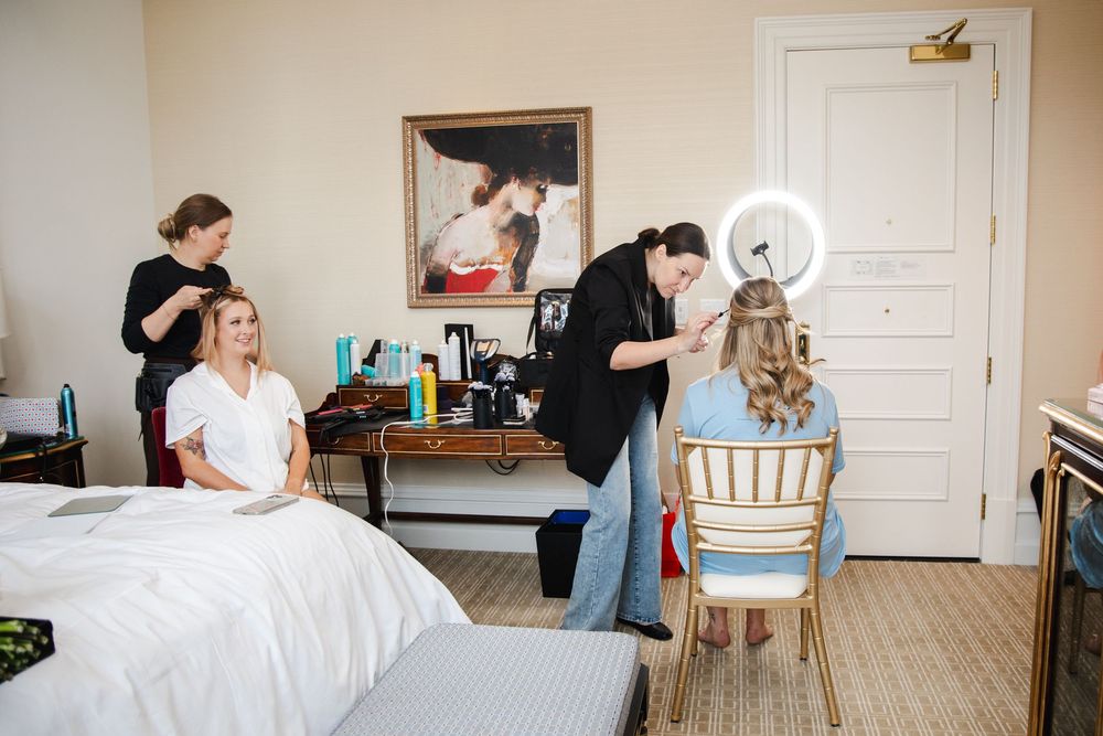 Bridal party getting ready in elegant room with makeup artist and hair stylist preparing two women.