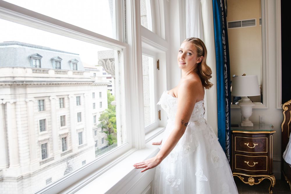 Bride in white wedding gown standing by large window with city view in elegant hotel room.
