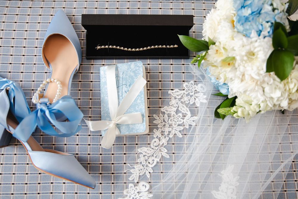 Blue bridal heels with ribbon, pearl necklace in box, gift box, lace veil, and white blue hydrangea bouquet flatlay.