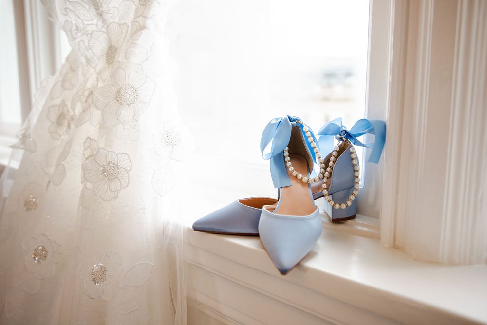 Light blue bridal heels with pearl accents and bow details on a windowsill beside a white lace wedding dress.