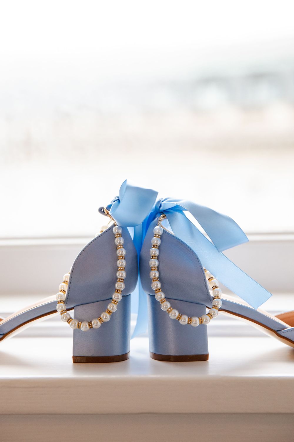 Light blue wedding heels adorned with pearl trim and a satin bow, displayed on a white windowsill.
