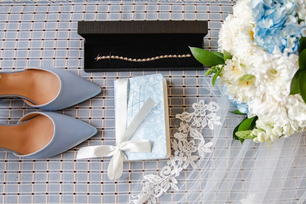 Elegant wedding flat lay with blue satin heels, pearl bracelet, clutch purse, lace veil, and blue hydrangea bouquet.