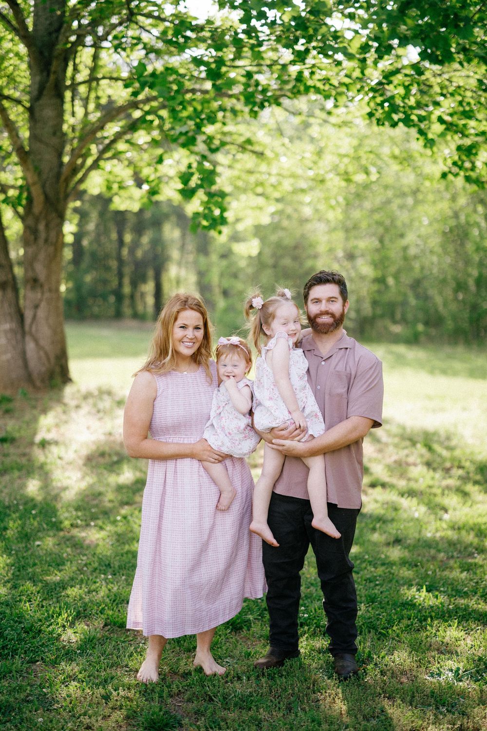 Family of four posing outdoors in green park, mother in pink dress holding baby, father with beard holding toddler girl.
