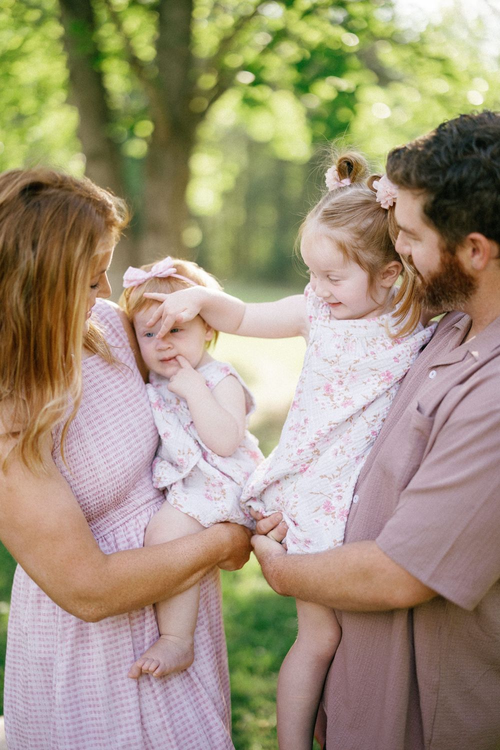 A young family with two small daughters in pink dresses enjoy a sunny outdoor moment among green trees.