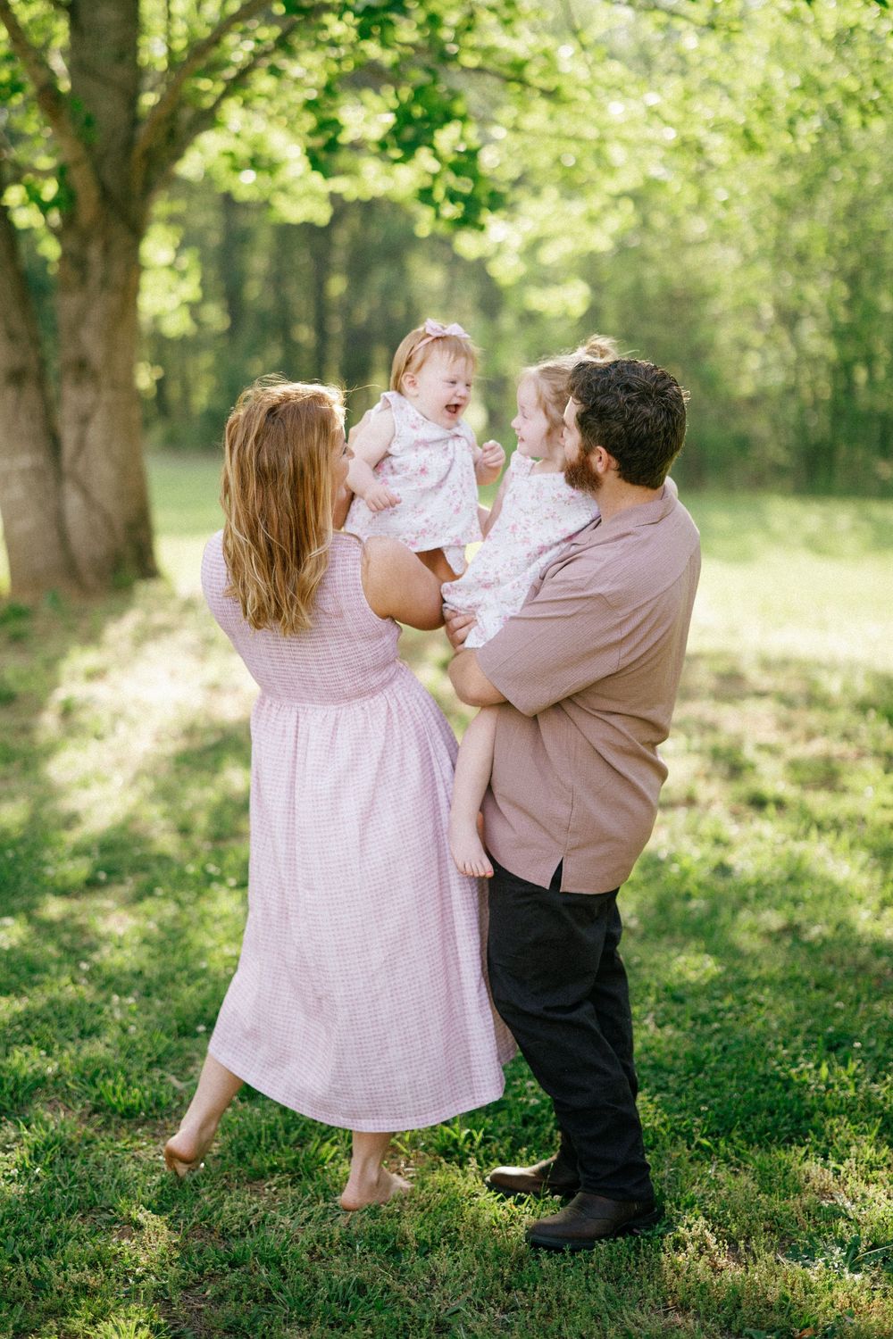 A happy family of four enjoys a sunny outdoor moment, with parents holding two young girls in a lush green park.