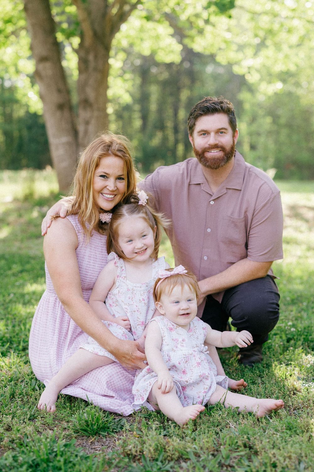 A smiling family of four poses outdoors on green grass with trees, featuring two young girls in floral dresses.