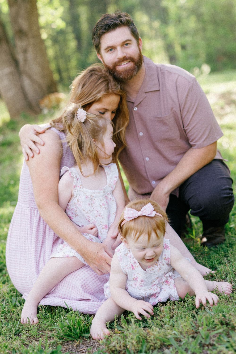 A bearded man and woman in pink sit on grass with their two young daughters during an outdoor family photo session.