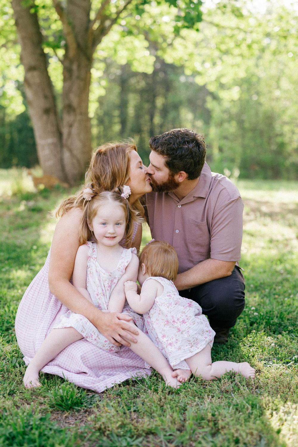 Family of four sitting on grass, parents sharing a kiss while two young daughters in pink dresses play together outdoors.