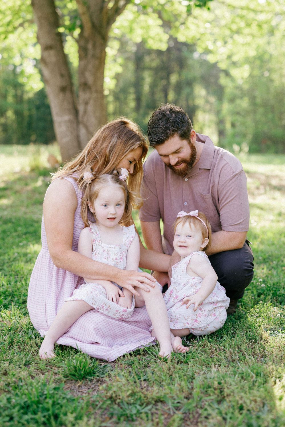 Happy family with two little girls in matching pink dresses sitting together on green grass under shady trees.