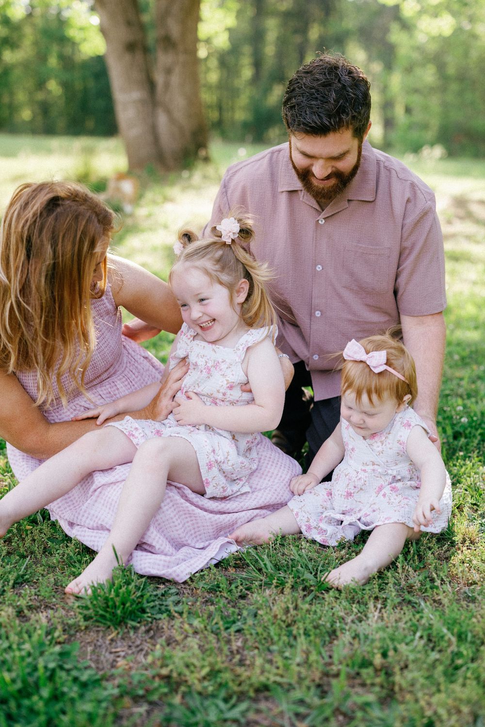Laughing toddler girl enjoys a tickle from mom while dad watches, family sitting on grass in sunny park.