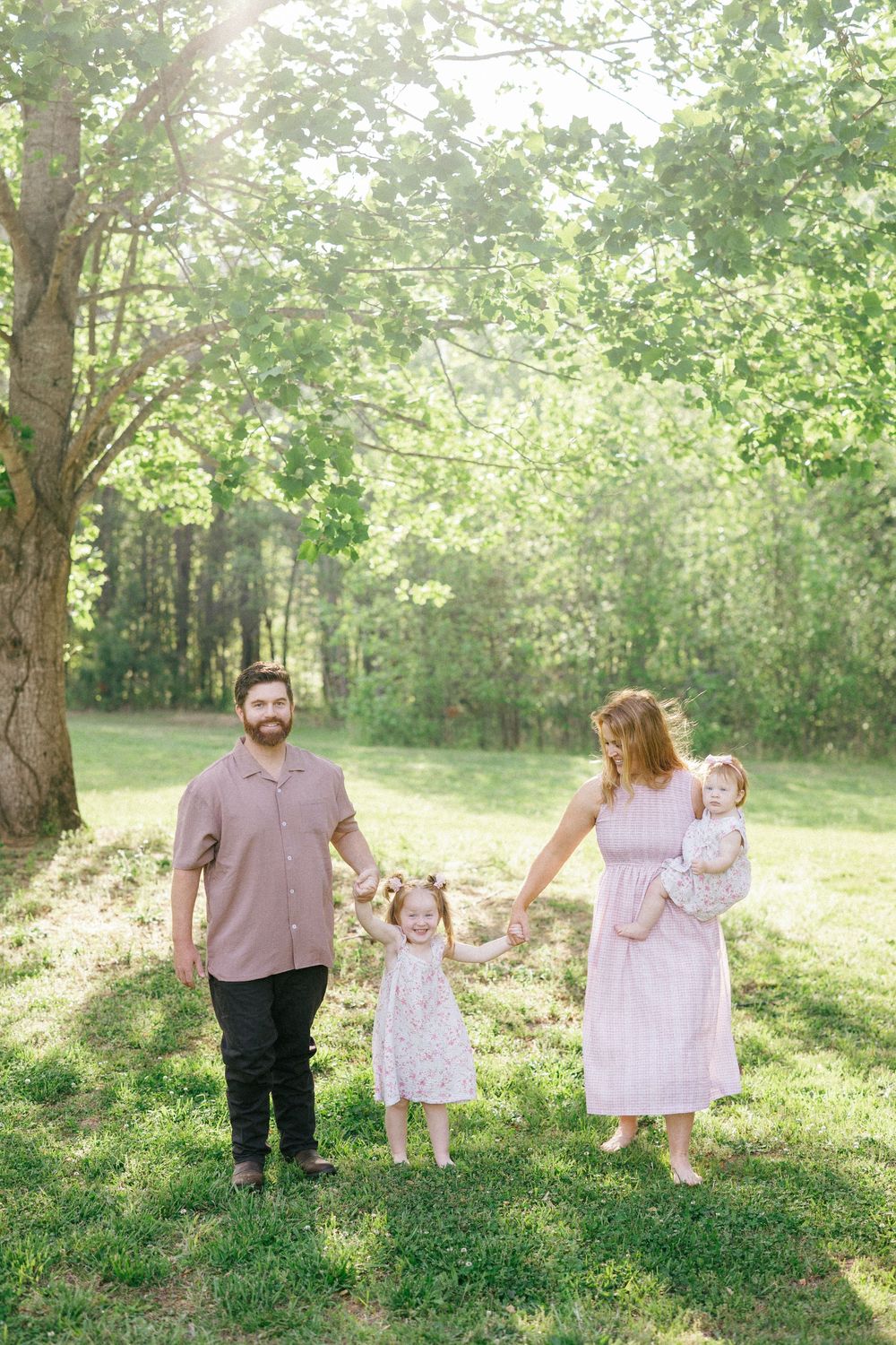 A smiling family of four walks outdoors in a sunlit green park, parents holding hands with a toddler girl while mom carries a baby.