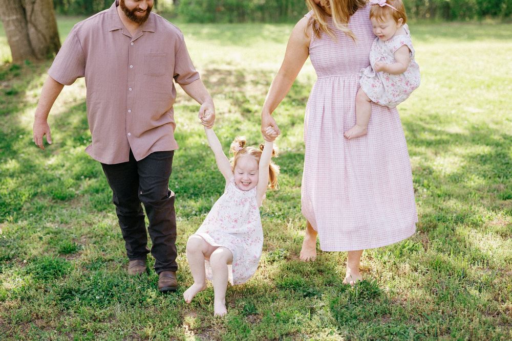 A family of four plays outdoors on green grass, parents swinging their toddler girl while holding a baby.