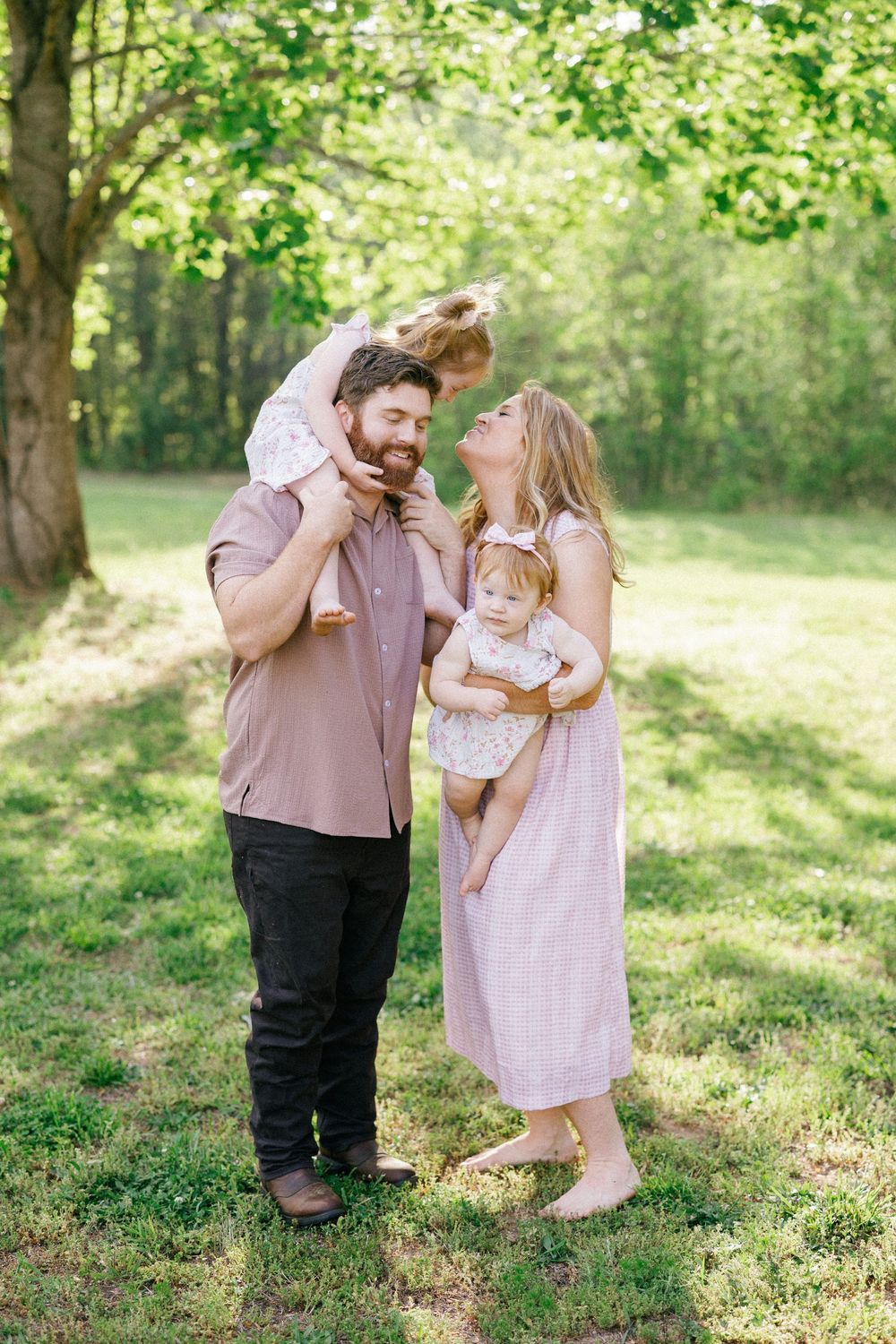 A happy family of four poses outdoors in a lush green park, with dad holding a toddler on his shoulders and mom cradling a baby.