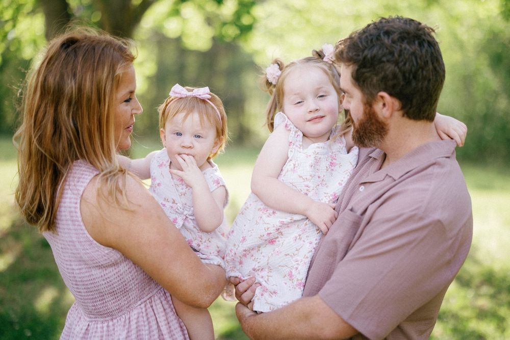 A happy family of four with two toddler girls in pink dresses posing outdoors in a lush green park setting.