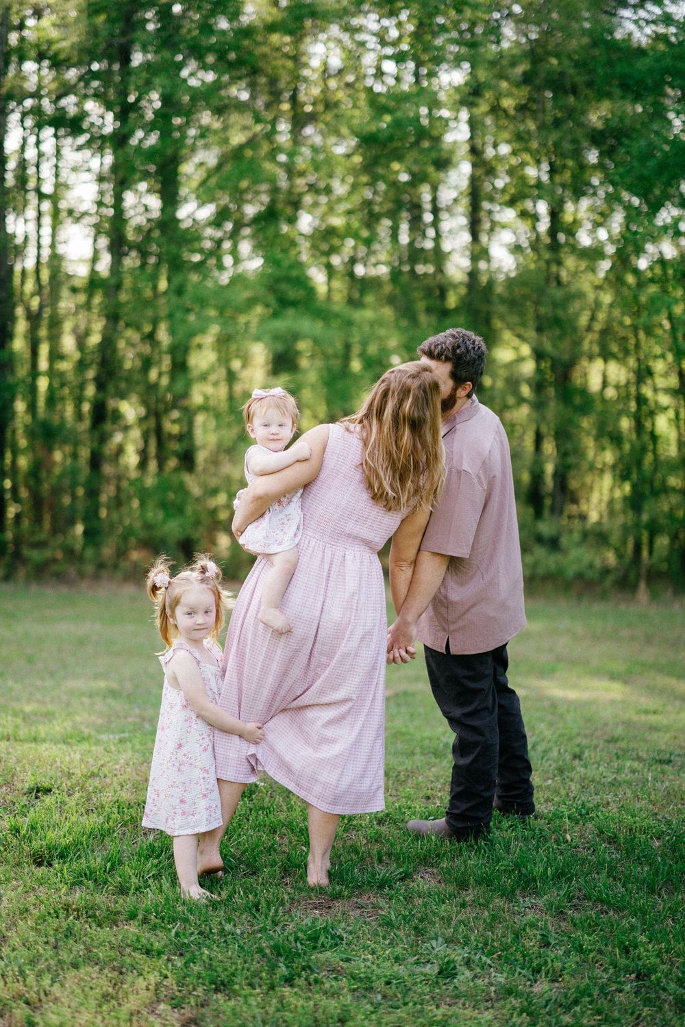Family of four standing in a green meadow, mother holding baby while kissing father, young daughter standing nearby.