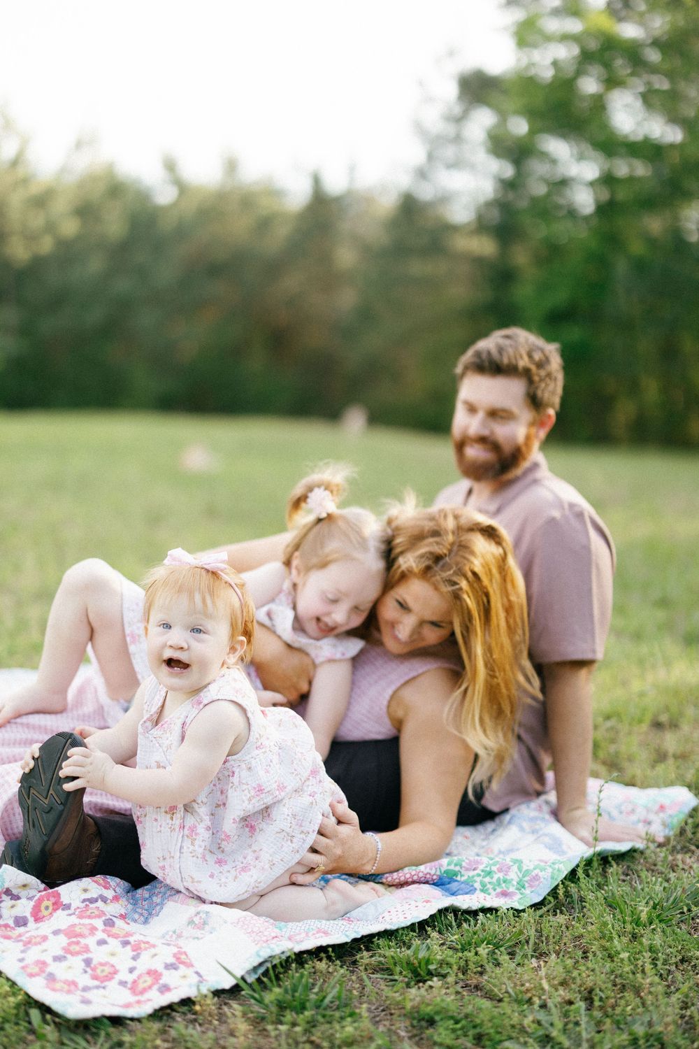 Happy family of four sitting on a blanket in a sunny green park, with two young girls and their smiling parents.