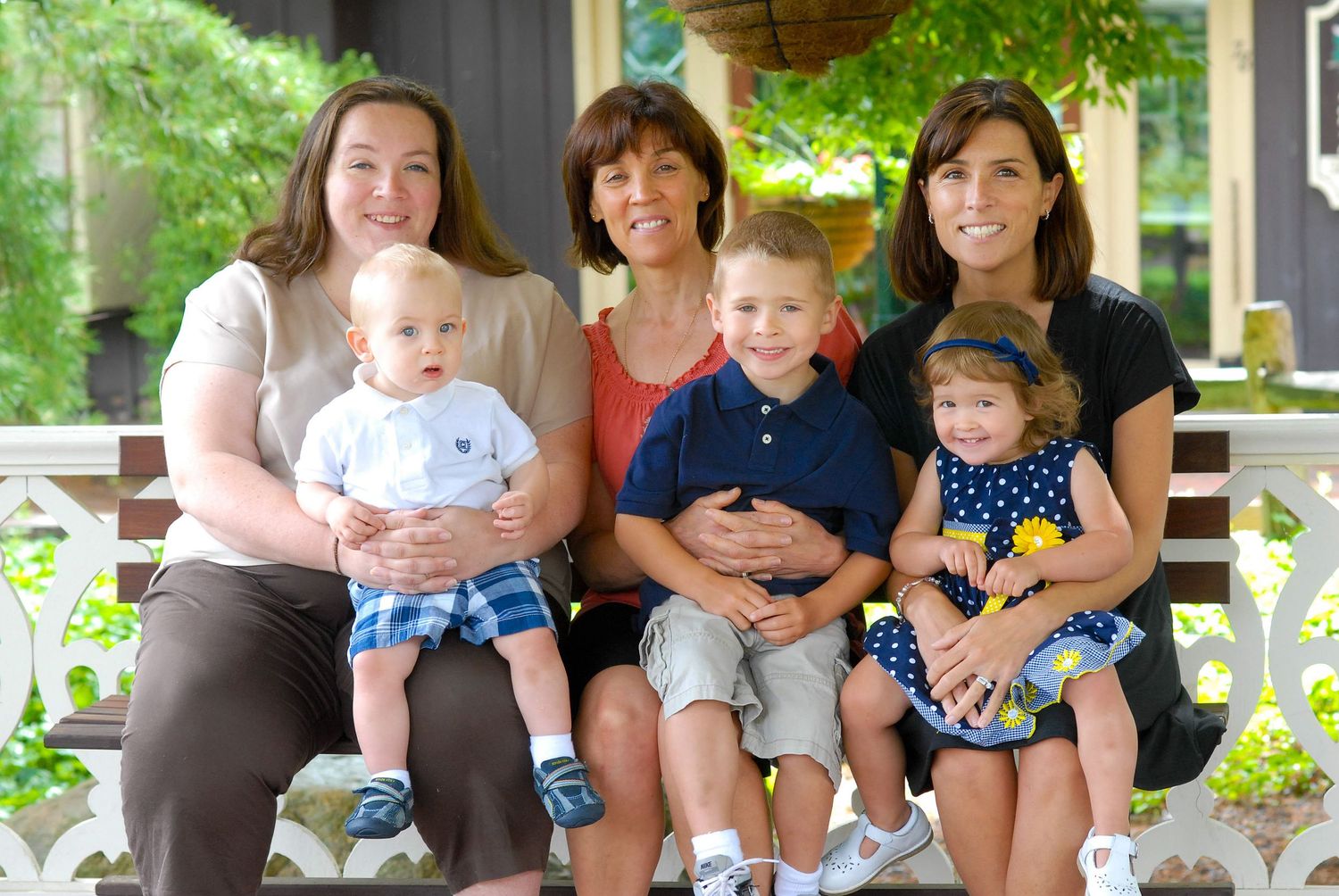 Three women and three young children smile together on a white bench on a porch for a family portrait.