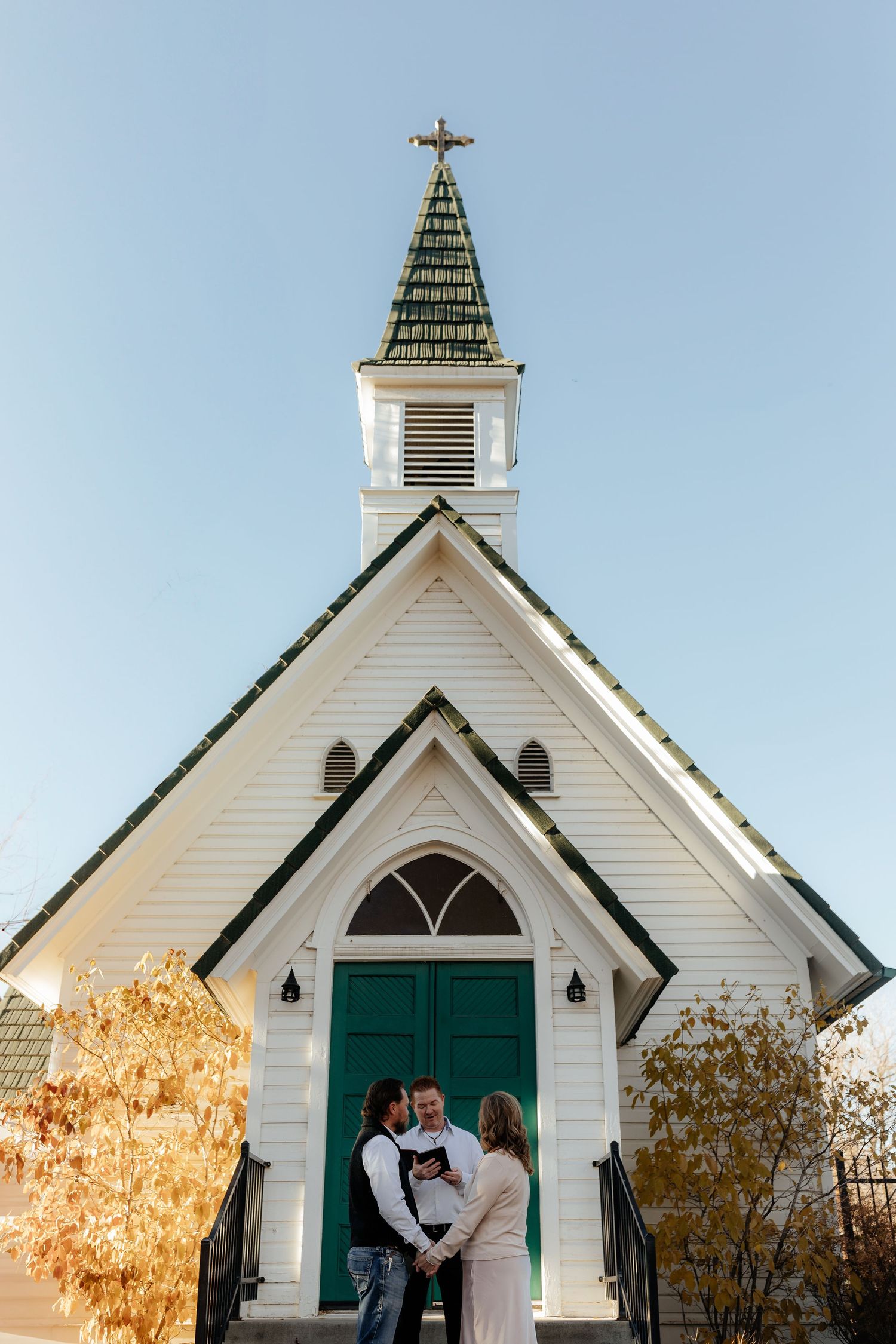 Couple sharing a kiss outside a charming white chapel with green doors and a cross-topped steeple in autumn.