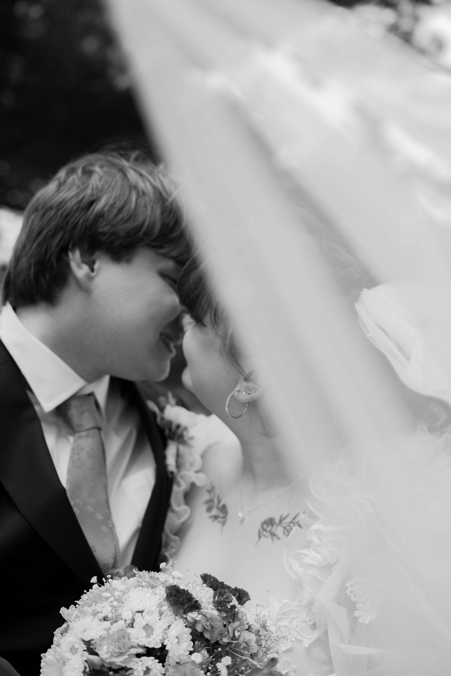 A black and white photo of a bride and groom sharing a kiss beneath a flowing wedding veil while holding a bouquet.