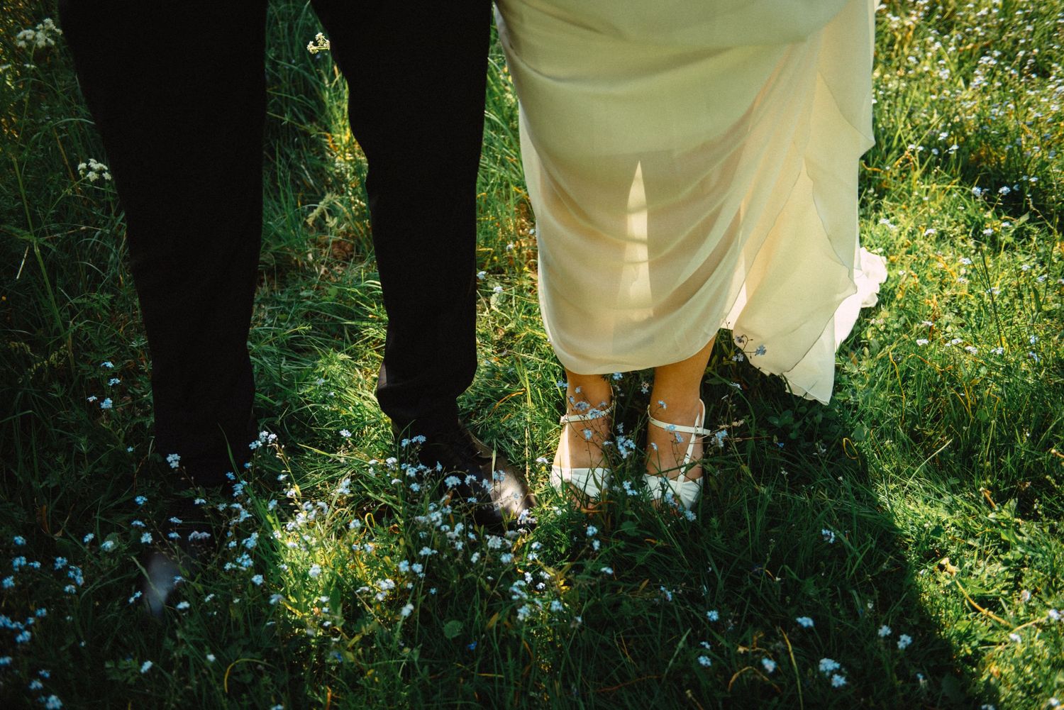 Bride in yellow dress and silver heels standing with groom in black suit on lush green grass with wildflowers.