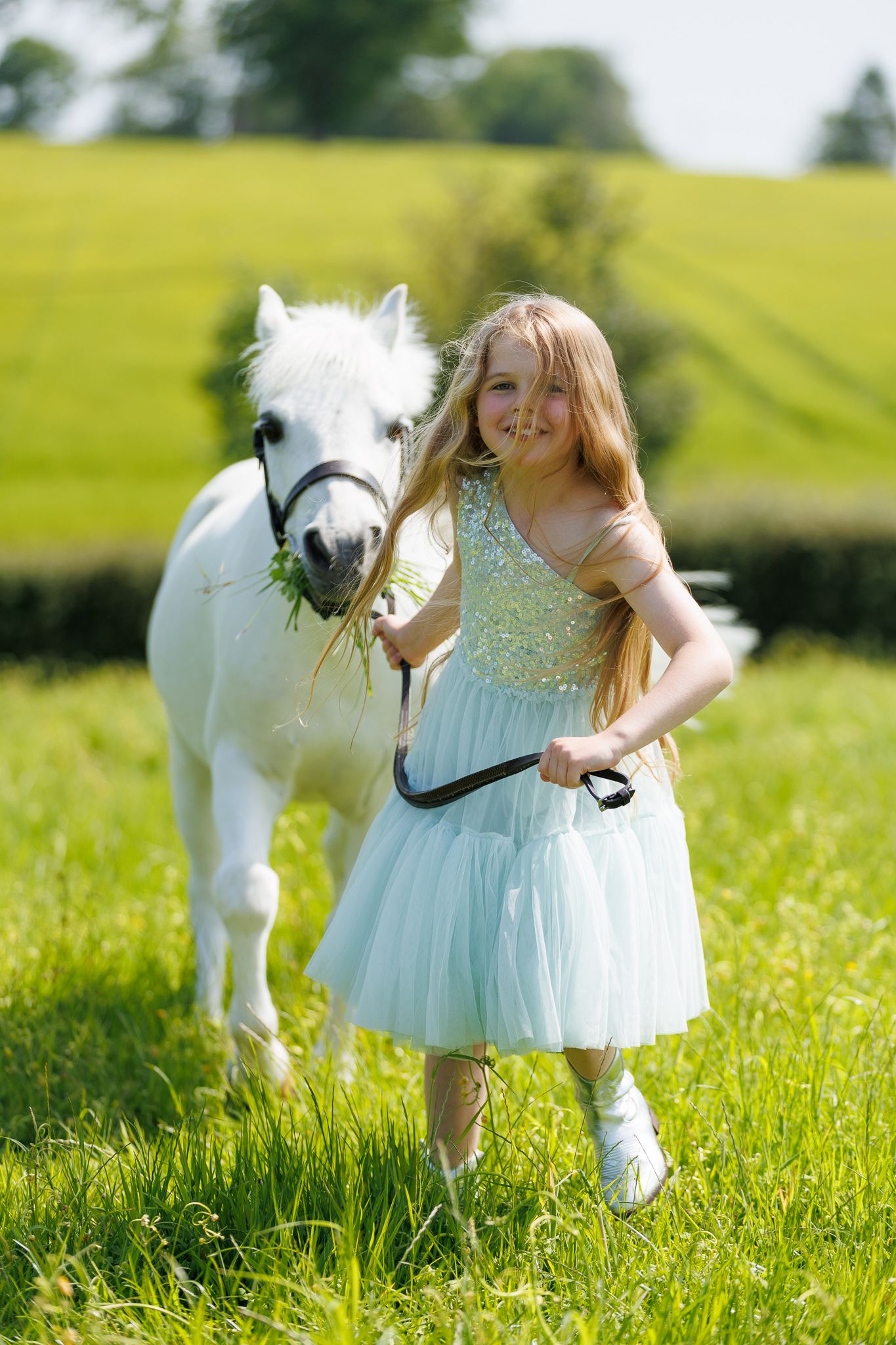 A beautiful bay horse wearing a black bridle stands next to someone in a white lace dress in an outdoor setting.