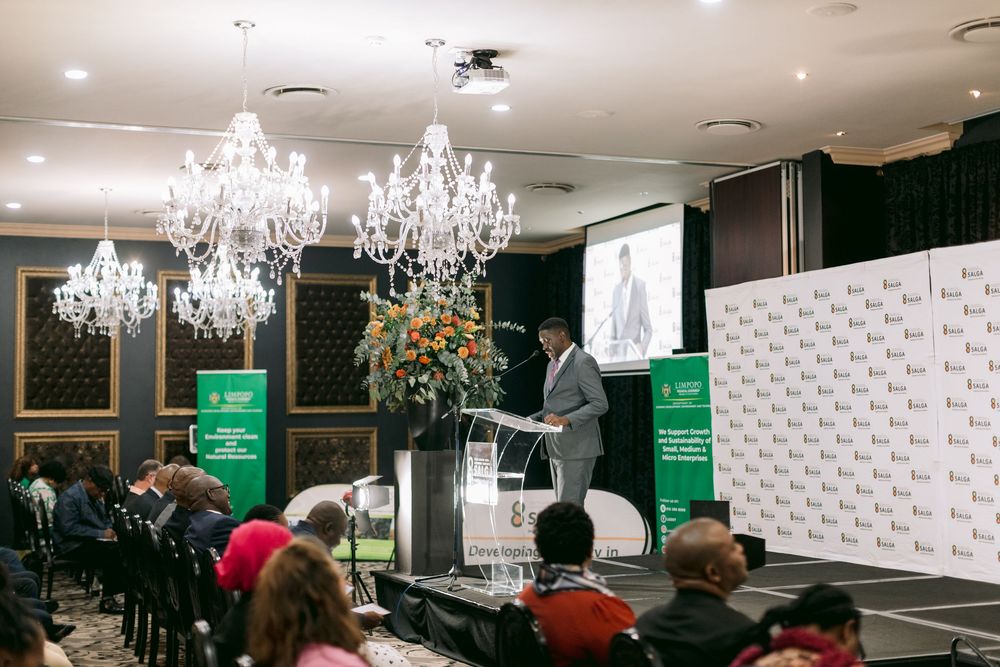 Speaker at glass podium addressing audience in elegant ballroom with chandeliers and green branded backdrop.