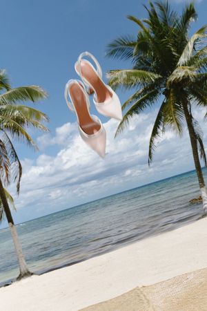White bridal heels tossed in air between palm trees on a sunny tropical beach.