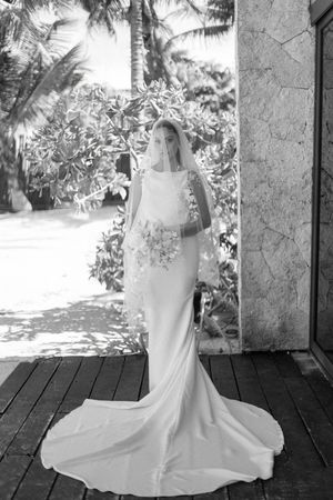 Elegant bride in fitted white gown and veil holds bouquet beside tropical stone building.