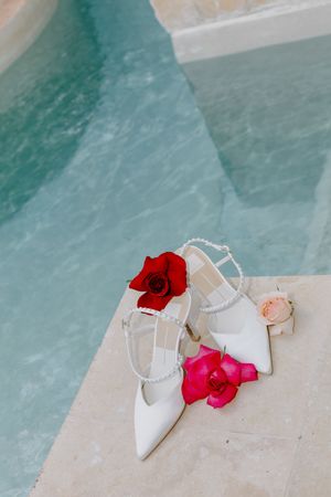 White bridal heels with pearl straps adorned with red and pink flowers by pool.