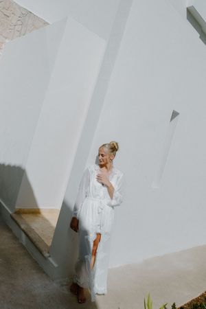 Blonde bride in white lace robe posing against a sunlit white wall outdoors.