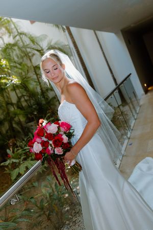 Blonde bride in white gown holding vibrant red and pink bouquet on staircase.
