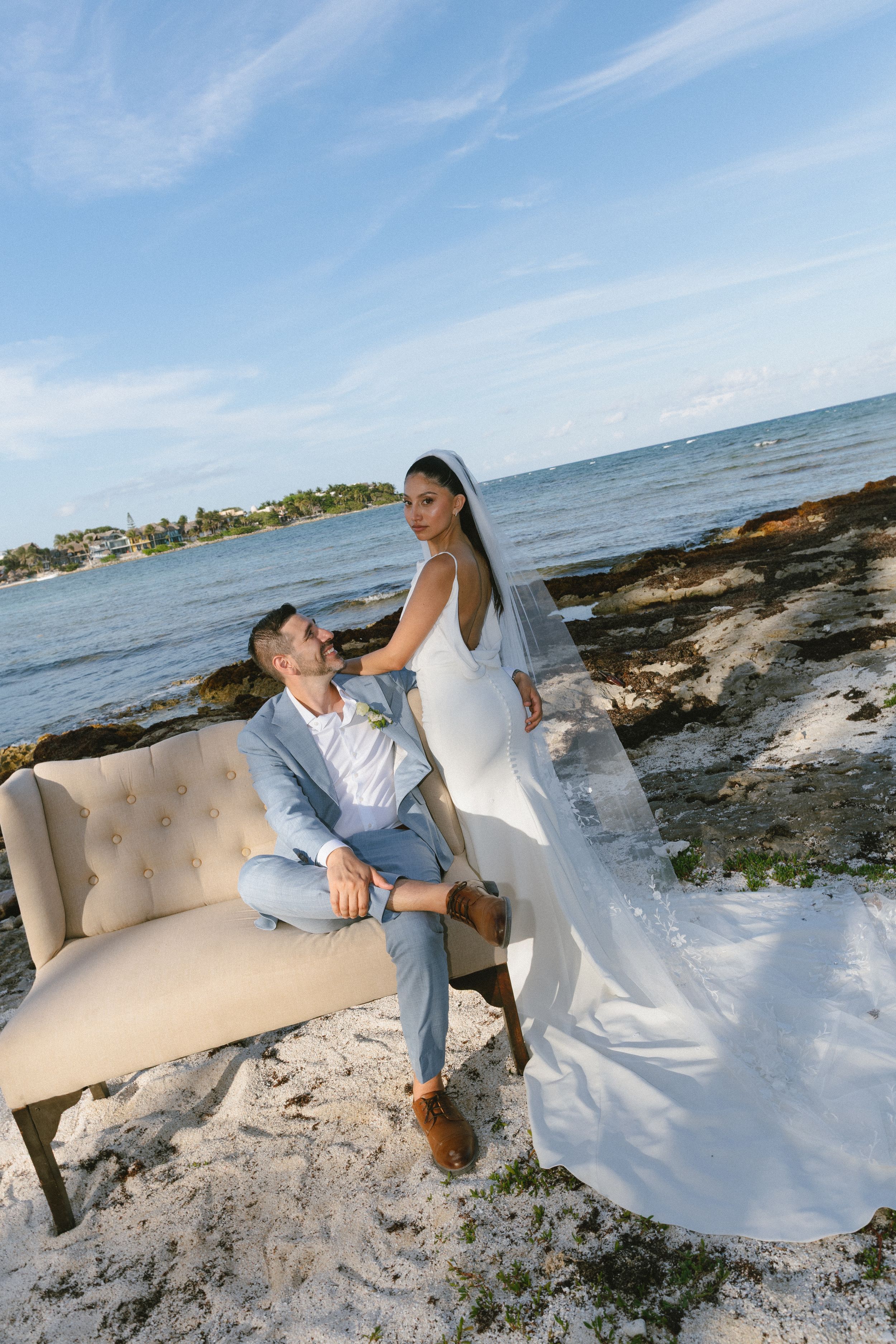 Bride in white gown and veil stands beside groom seated on beach sofa with ocean backdrop.