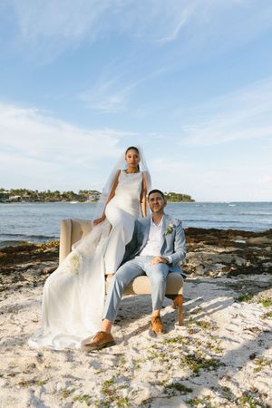 Bride and groom posing together on a rocky beach with ocean and blue sky in background.