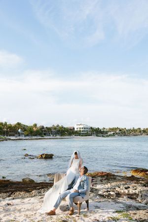 Bride and groom posing on rocky Caribbean beach shoreline with ocean and blue sky backdrop.