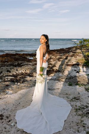 Elegant bride in white fitted gown holding bouquet standing on rocky beach by the ocean.