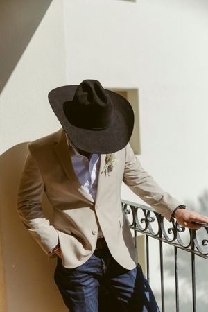 A groom in a tan blazer and black cowboy hat leans against a railing in sunlight.