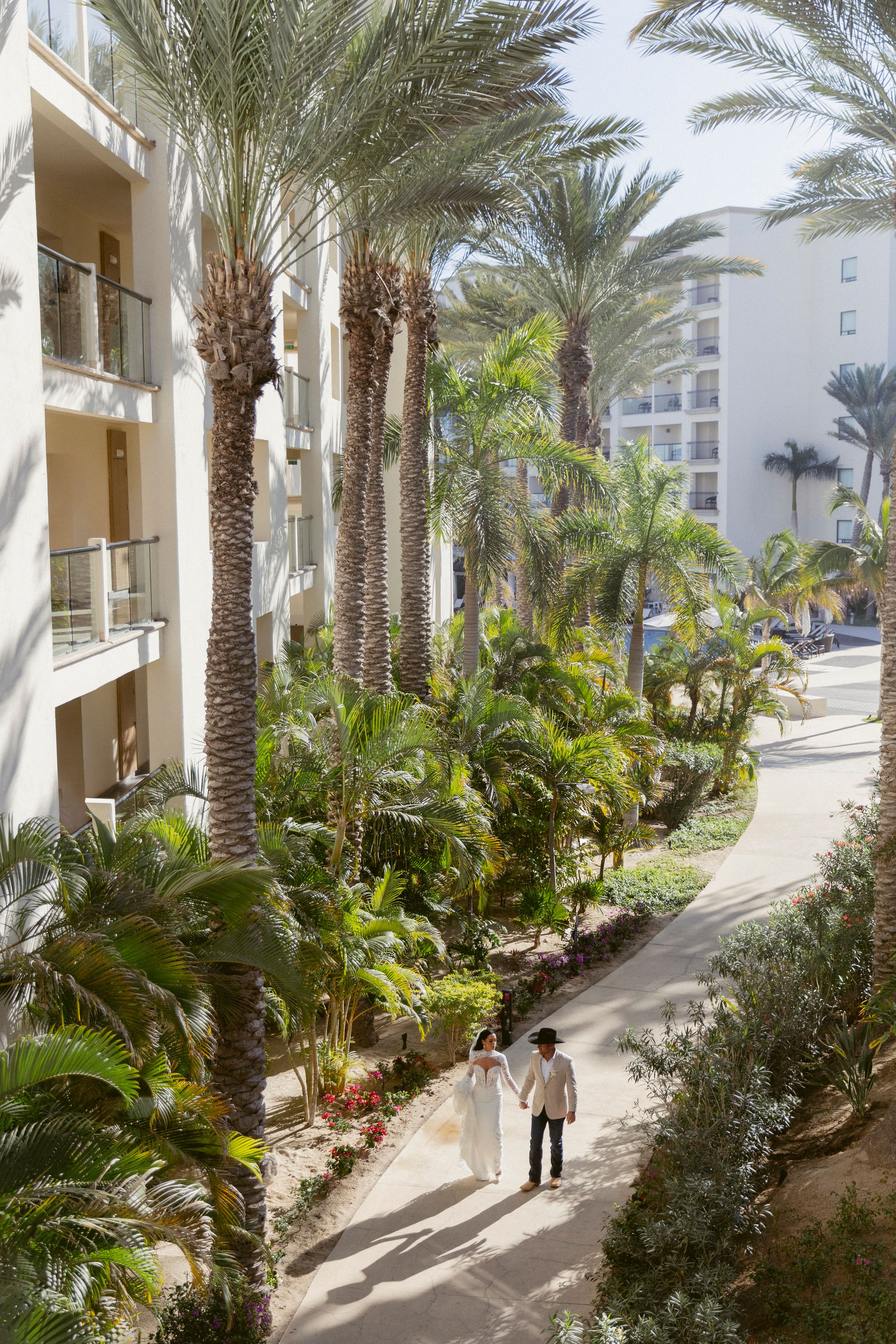 A newlywed couple walks hand in hand along a palm tree-lined resort pathway.