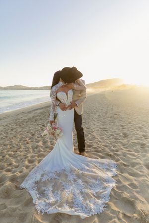 A bride and groom share a kiss on a sandy beach at golden sunset.