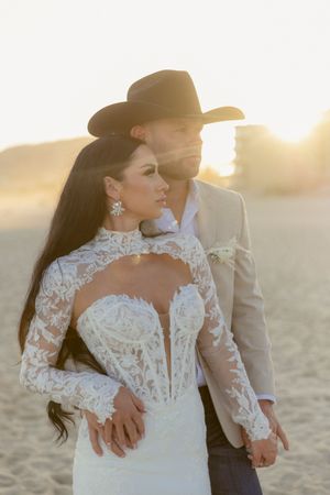 A bride in lace gown and groom in cowboy hat embrace on a sunlit beach.