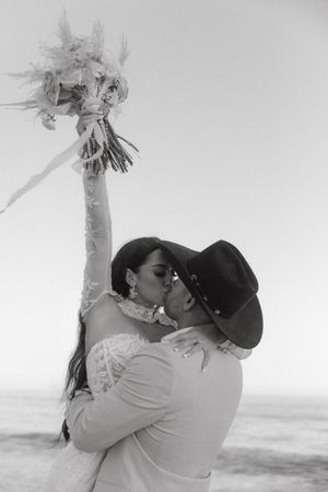 A bride and groom kiss on the beach as the bride raises her floral bouquet skyward.