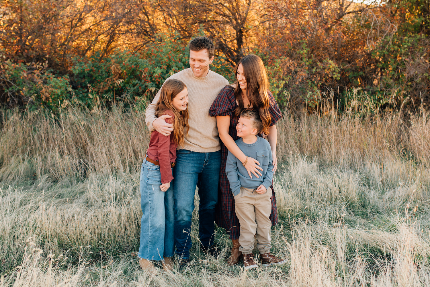 Family of four posing together in golden autumn field, parents and two young children smiling warmly at sunset.