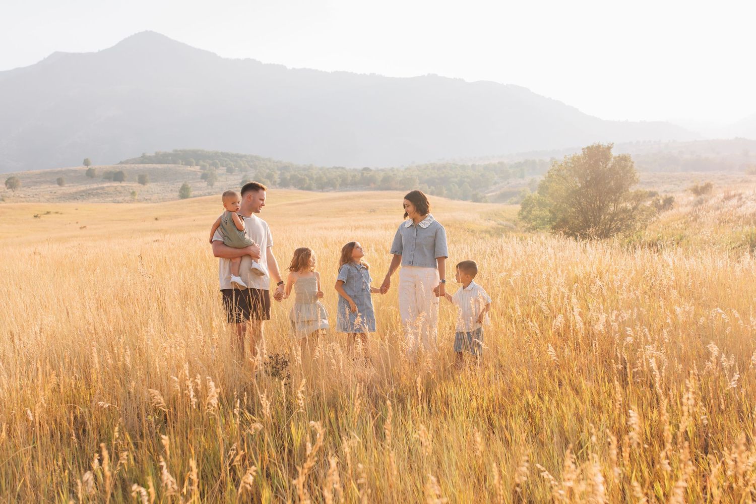 Family of six standing in golden field with mountain backdrop in warm sunlight.