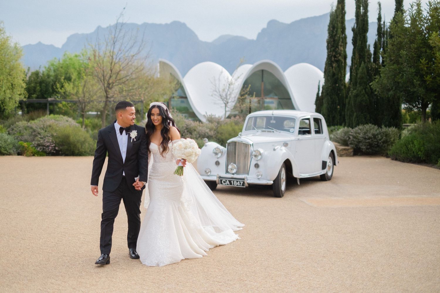 Bride and groom walk hand in hand by vintage white car with mountain backdrop at venue.