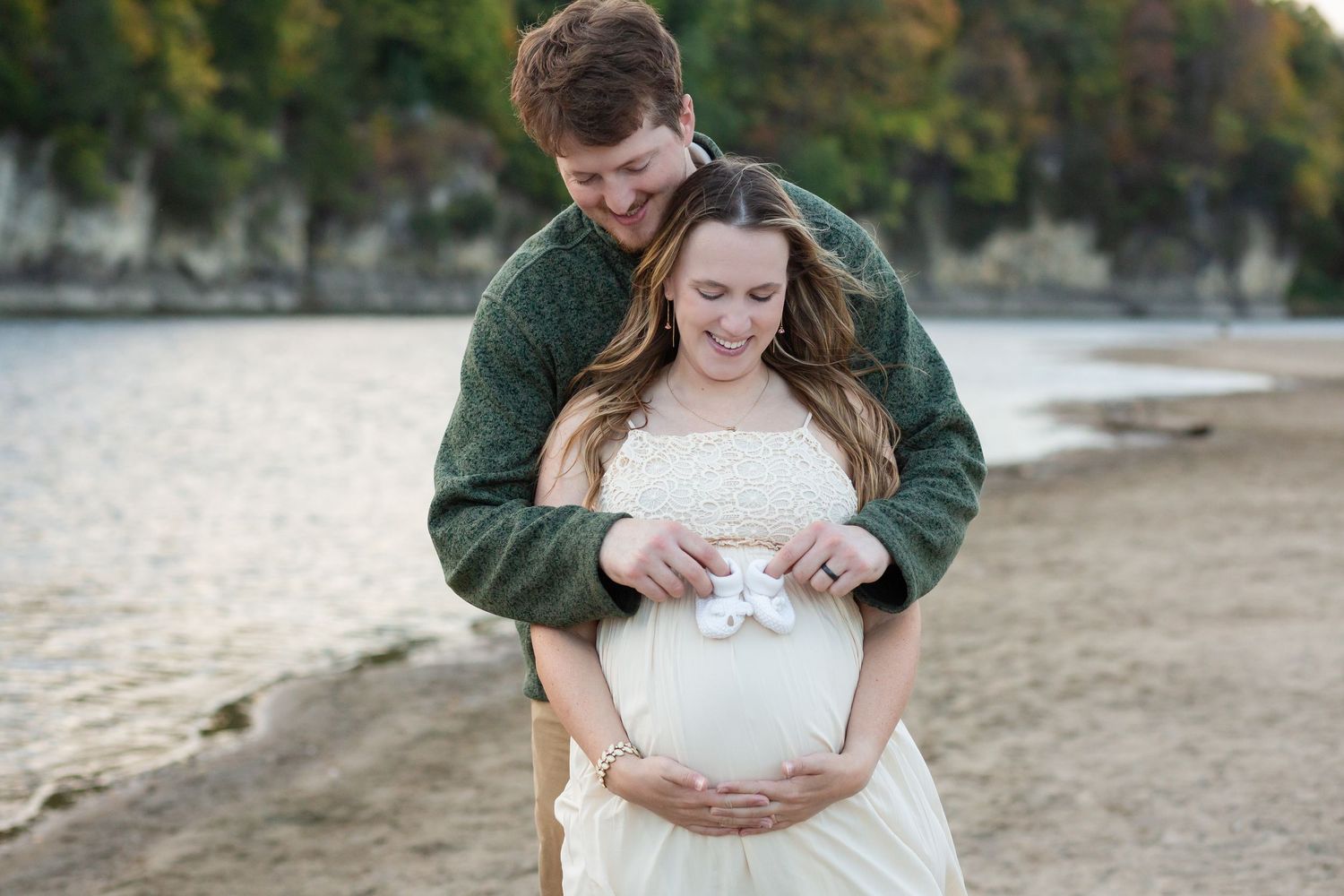 A couple poses on a beach, man embracing pregnant woman in white dress, both smiling at tiny baby shoes on her belly.