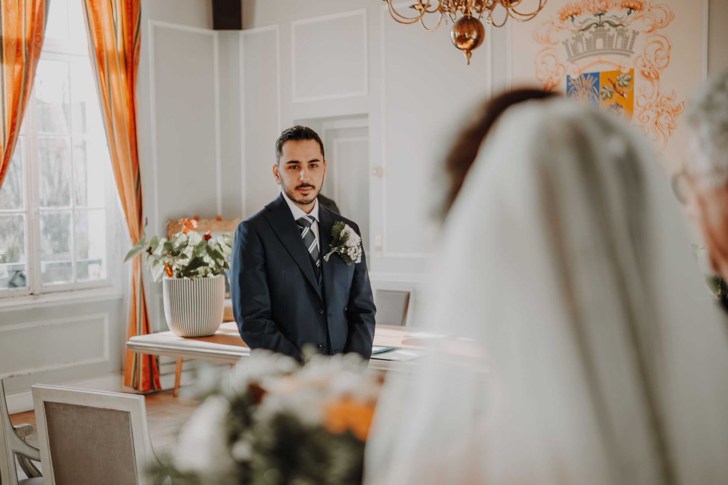 Groom in navy suit watches bride approach during elegant indoor wedding ceremony with floral arrangements.