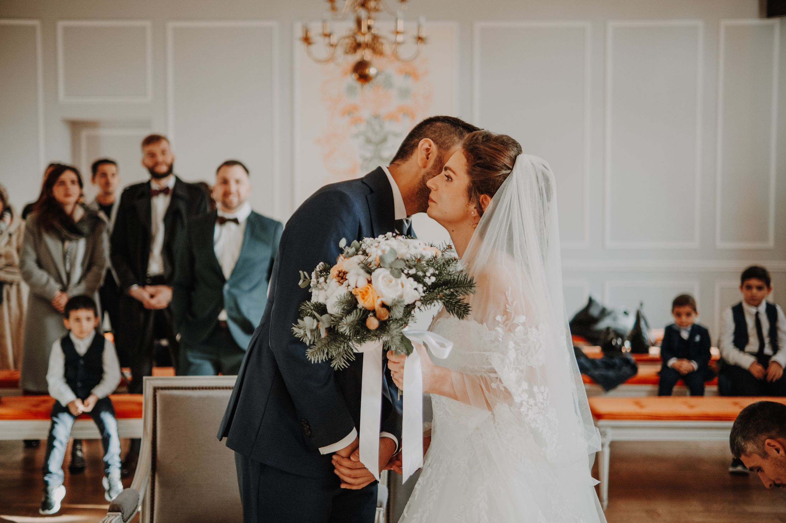 Bride and groom share first kiss at wedding ceremony in elegant white chapel with guests watching.