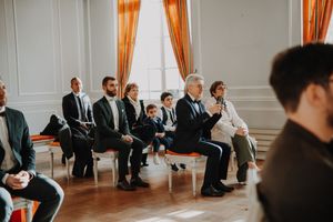 Guests seated in elegant room with orange curtains during a formal ceremony or wedding event.