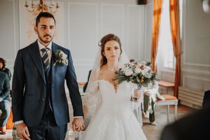 Bride and groom holding hands at wedding ceremony, bride in lace gown holding bouquet, groom in navy suit.